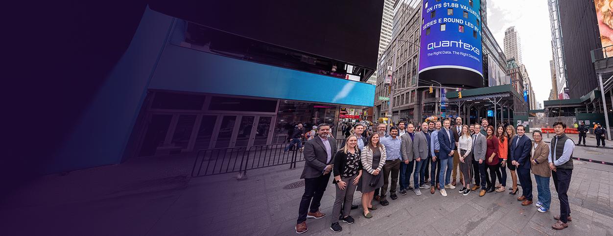 A group of people stands together in a cityscape, with a large digital billboard in the background displaying text.