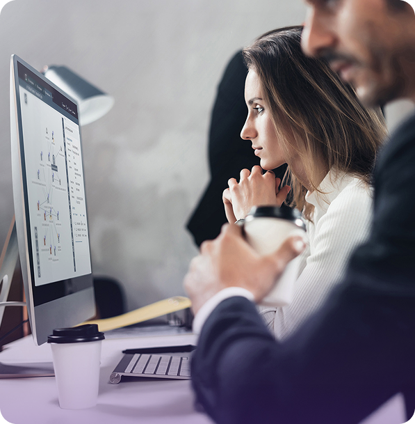 Woman focused on a computer screen displaying diagrams, while a man in the foreground holds a coffee cup.